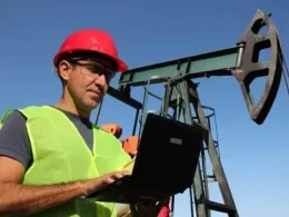 A man in a hard hat on a laptop in front of an oil derrick