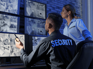 A male and female security security guards viewing CCTV monitors in a dark room.