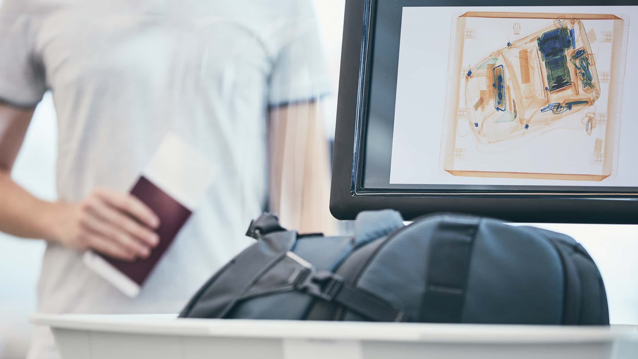 A bag in a tray about to go through and airport scanner as a man stands behind holding his passport.