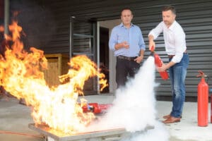 A man instructing another man on how to use a fire extinguisher as he operates it on a small controlled file.