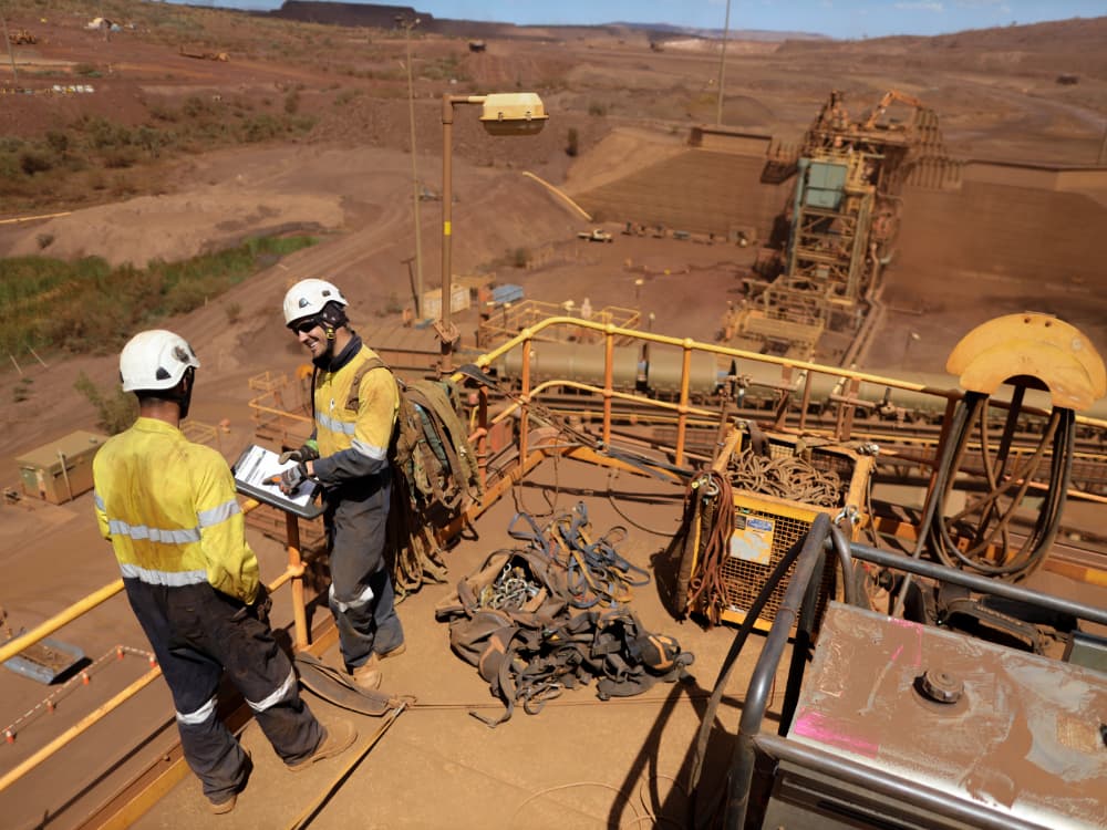 Two miners standing on a high platform in the sun over a red dirt mining site.