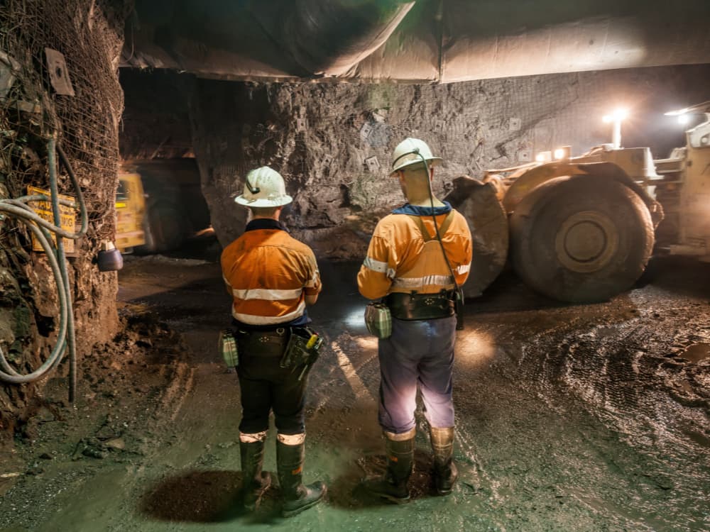 Two miners in high vis facing a large mining equipment in an underground mine.